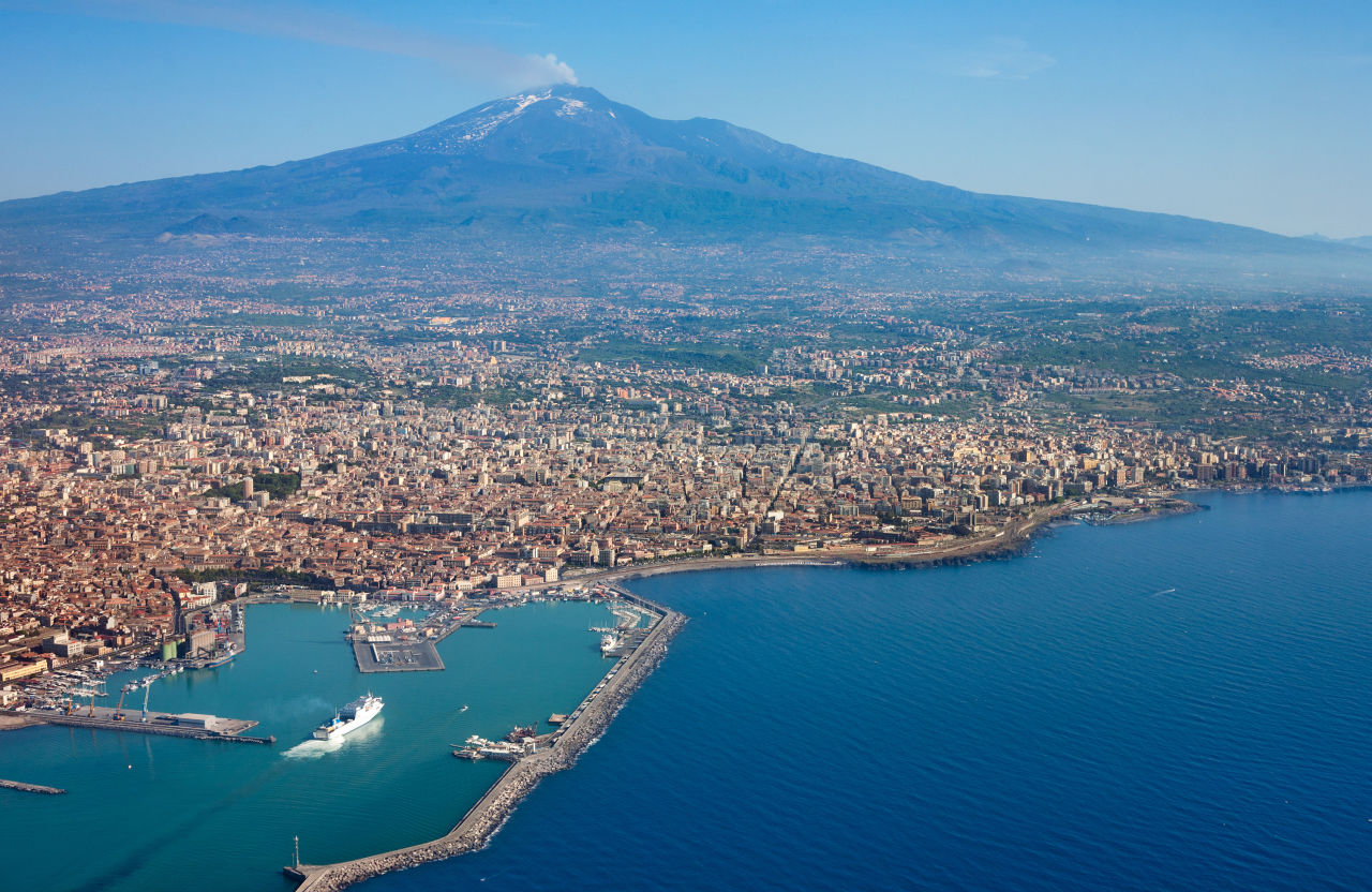 Panorama Katanii z lotu ptaka, ukazująca tętniący życiem port, rozległą zabudowę miejską i majestatyczny wulkan Etna w tle.