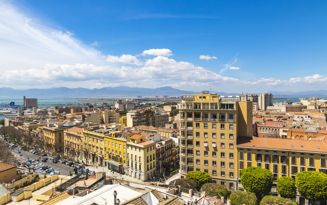 Rozległa panorama Cagliari ukazująca urok historycznych budynków, nowoczesnej zabudowy i portu, z majestatycznymi górami w tle pod błękitnym niebem.
