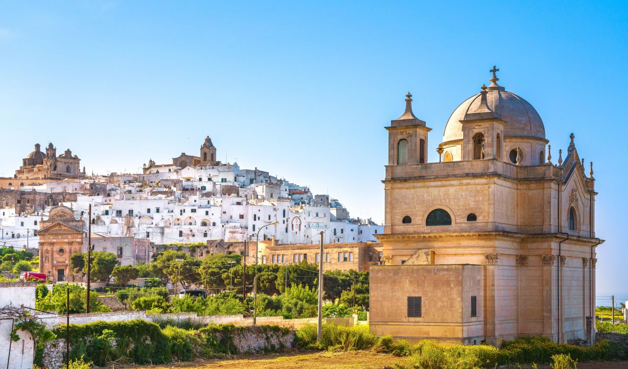 Blick von Weitem auf das Panorama der weißen Stadt Ostuni mit mittelalterlichem Kirchturm im Vordergrund