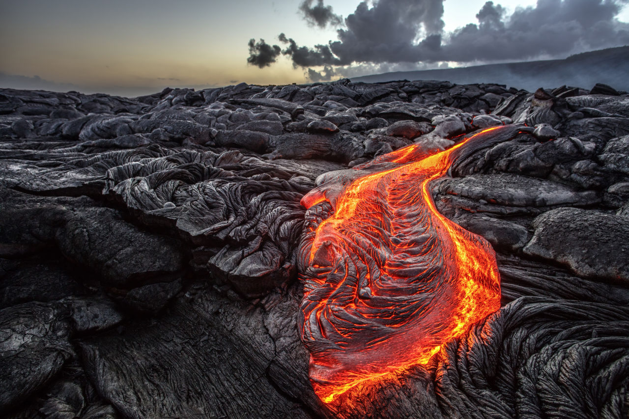 Glowing orange lava flowing through hardened black basalt during a fissure eruption on Iceland’s Reykjanes Peninsula at sunset