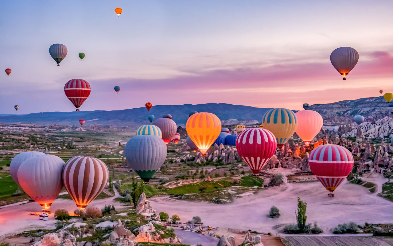 Flüge mit dem Heißluftballon sind im Kappadokien Urlaub sehr beliebt – hier ein schönes Panorama mit dem Blick auf eine malerische Stadt und die Ballons am Himmel darüber