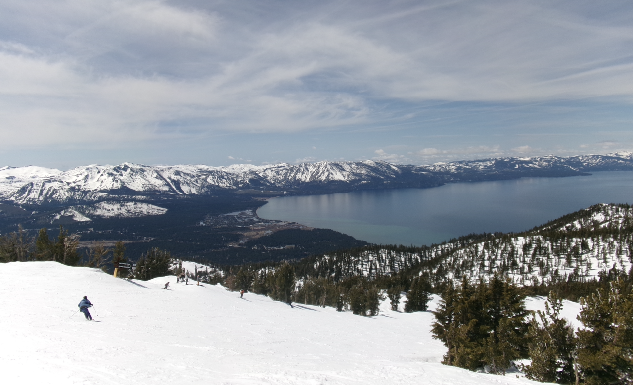 Skiers descending a snow-covered slope above Lake Tahoe with the Sierra Nevada mountains and blue lake visible in the background.