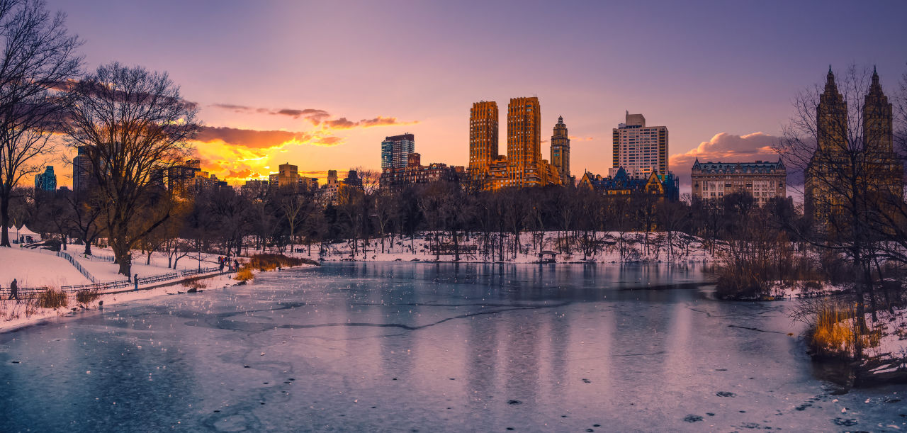 Snow-covered Central Park at sunset with frozen lake and Upper West Side skyline glowing in golden winter light, New York City