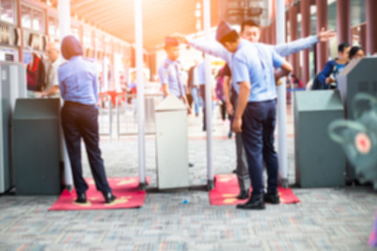 Airport security screening with passengers walking through metal detectors and security officers checking travelers at departure terminal.