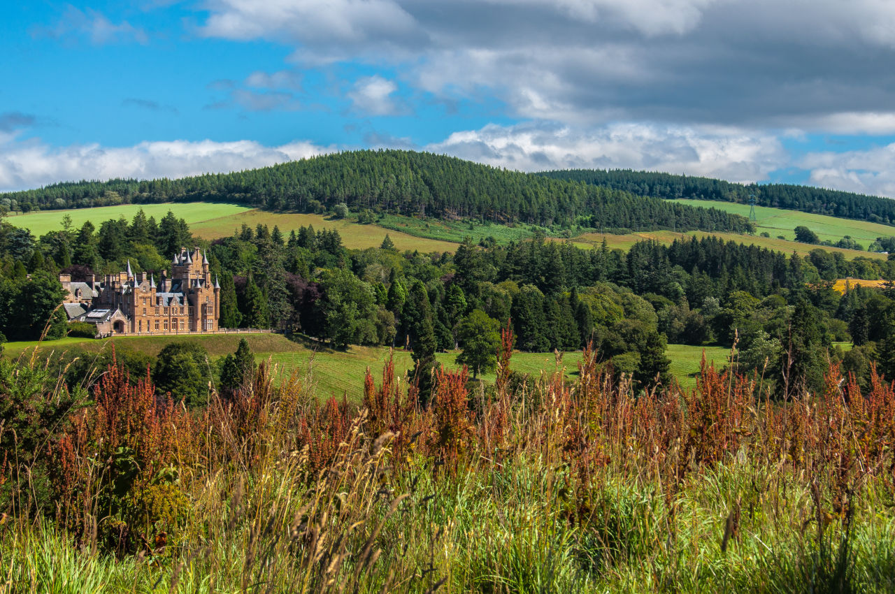 Ardross Castle surrounded by rolling green hills and forested countryside in the Scottish Highlands under a partly cloudy sky.