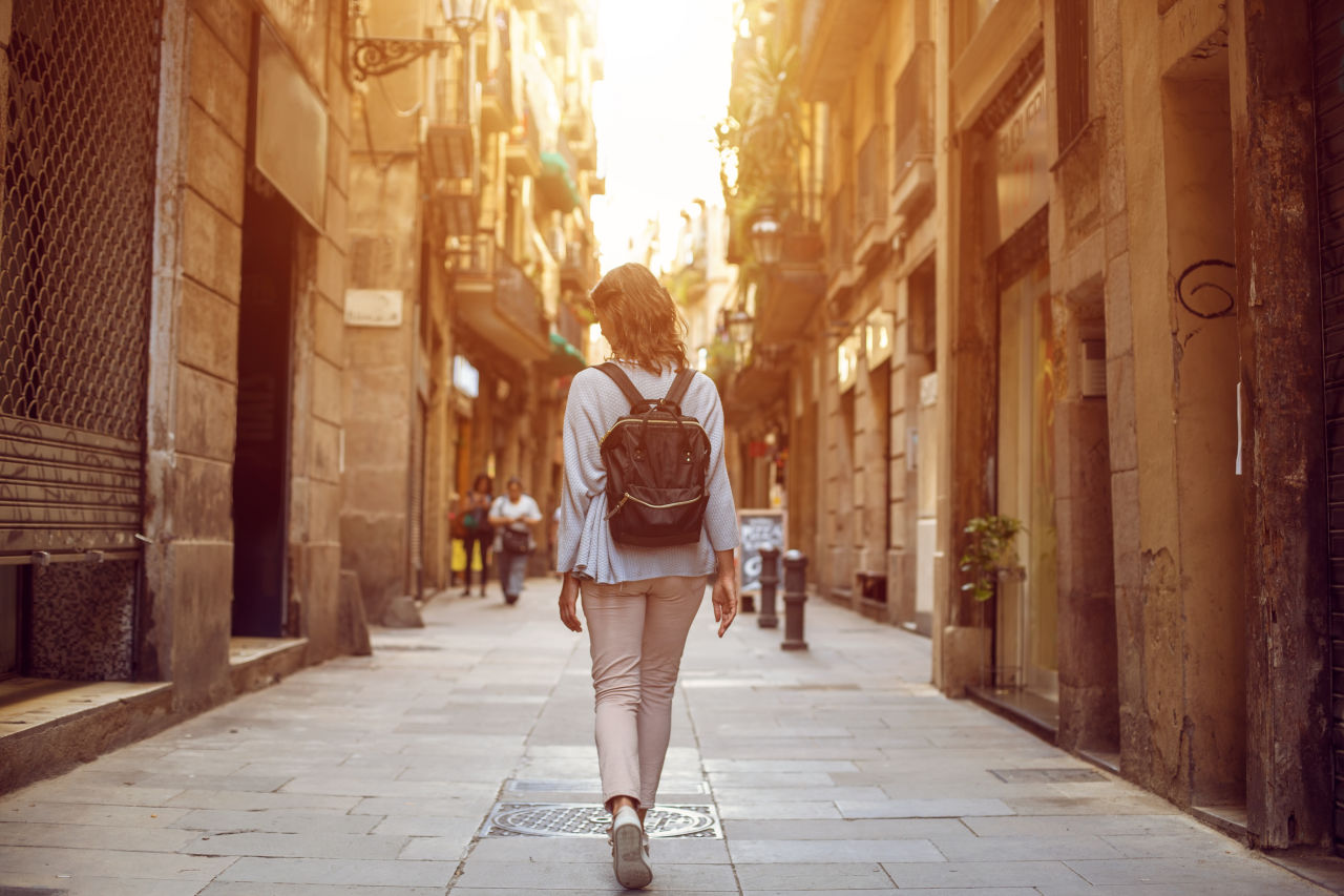 8 Destinations Travel Experts Say to Skip This Summer - Woman walking down a narrow European street at golden hour with backpack
