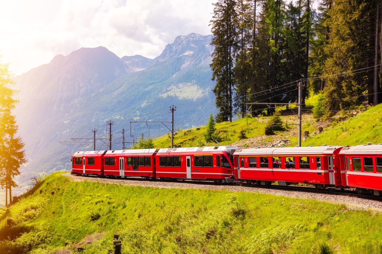   Bright red passenger train winding through the Swiss Alps with green hills, pine forests, and dramatic mountain scenery in summer sunlight.