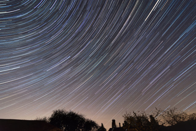 Multiple bright meteor trails streak across a blue-grey night sky above the dark silhouette of a bare tree