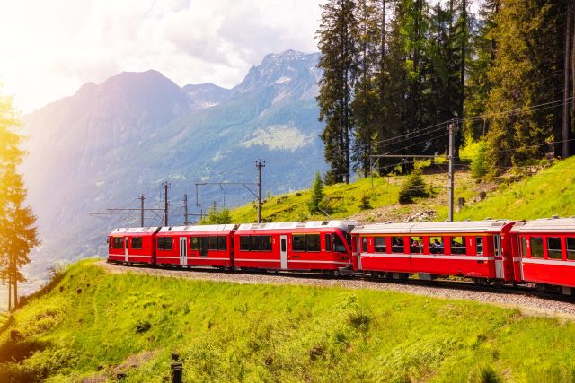   Bright red passenger train winding through the Swiss Alps with green hills, pine forests, and dramatic mountain scenery in summer sunlight.