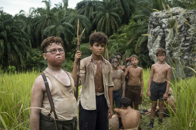 A group of young boys from the series stands in a tall grassy field with dense tropical palm trees in the background. In the foreground, a boy wearing round glasses, a tank top, and suspenders stands alongside a boy holding a sharpened wooden spear, both looking off-camera with serious expressions.