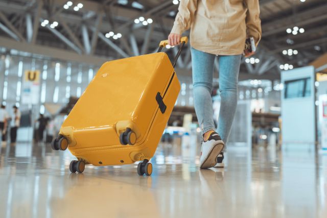 Airline Baggage Fees Are Going Up - A traveler wheels a bright yellow hard-shell suitcase through a modern airport terminal, passport in hand. Only the lower half of the person is visible, with blurred passengers and terminal signage in the background.