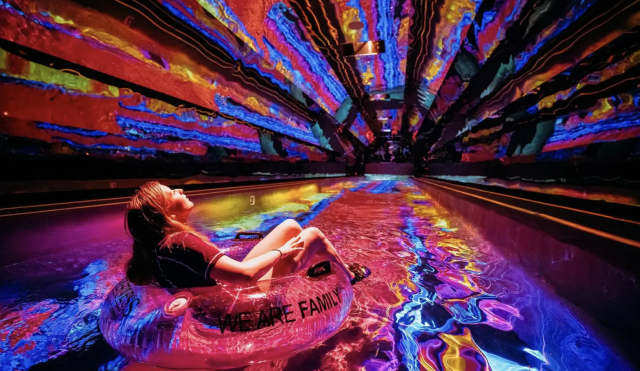 Guest floating on a tube through a neon-lit lazy river with colorful light projections covering the walls and ceiling