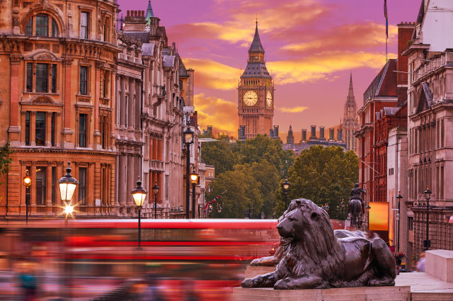 A cinematic, daylight establishing shot of central London looking down Whitehall from Trafalgar Square. Pedestrians and iconic red buses fill the streets, with the distinct silhouette of Big Ben rising in the background—a grounded, gritty visual framing a classic Jack Ryan political thriller setting.