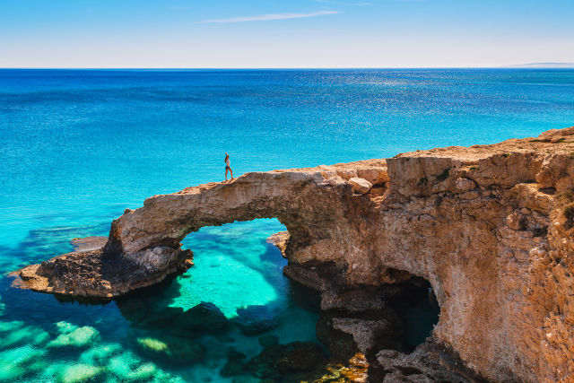Cyprus Drops to Level 1 Advisory as Mediterranean Travel Reopens - Person standing on a natural rock arch bridge over turquoise water at Cape Greko, Cyprus