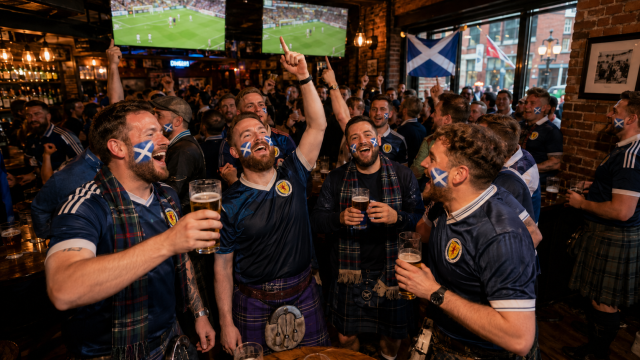 Scottish soccer fans wearing team shirts, tartan scarves, and kilts raise their beers and cheer inside a crowded bar.