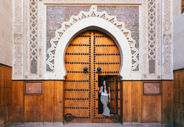 A woman standing in a carved wooden Moroccan doorway with intricate tilework, typical of Fez's historic medina architecture.