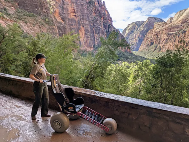 A female park ranger standing beside a red Extreme Motus all-terrain wheelchair on a paved canyon overlook path with sweeping views of Zion's rock walls behind her.