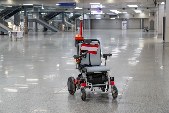 An autonomous wheelchair stands in a spacious airport terminal with sensors and a warning light mounted above the seat.