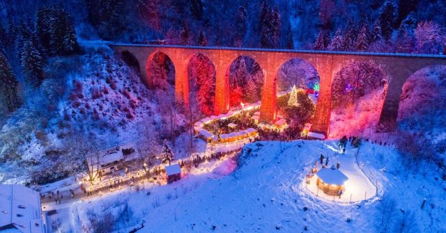 Aerial view of the snowy Ravenna Gorge Christmas Market beneath an illuminated railway viaduct in the Black Forest.