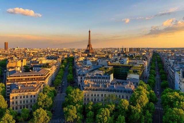 Paris skyline at sunset with the Eiffel Tower rising above tree-lined avenues and Haussmann rooftops, seen from the Arc de Triomphe