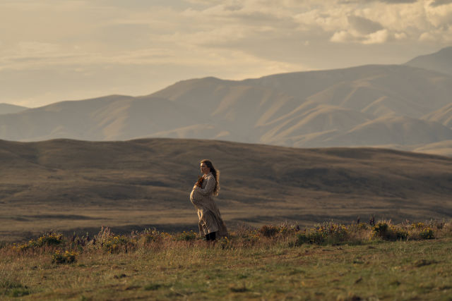 lorence Pugh as a pregnant Cathy Ames stands on a windswept hillside of wildflowers with golden hills behind her in Netflix's East of Eden, filmed in New Zealand